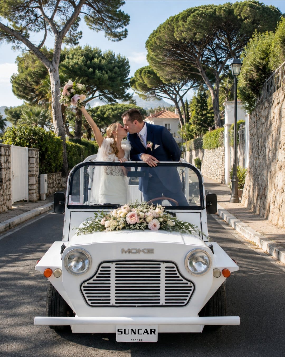 Mariés en Moke électrique blanche décorée de fleurs, sur une route de la Côte d'Azur