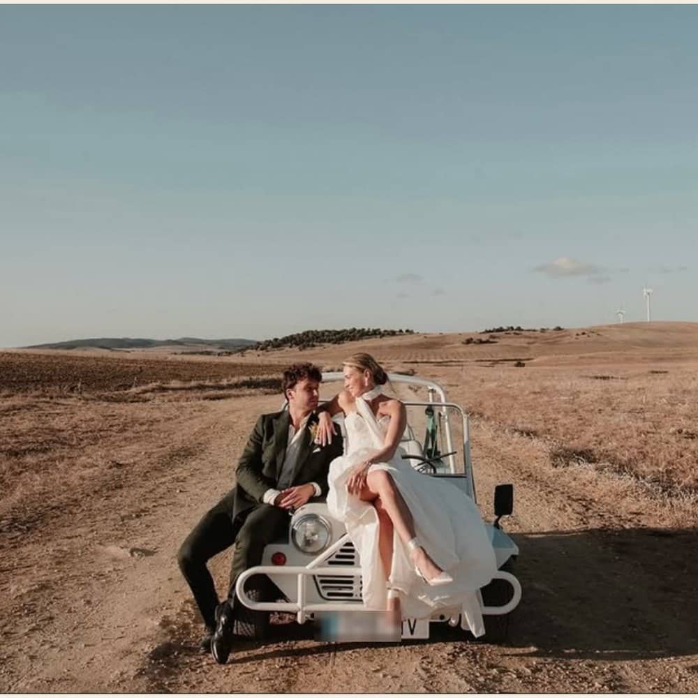 Couple de mariés assis sur le capot d'une Moke électrique blanche en pleine campagne