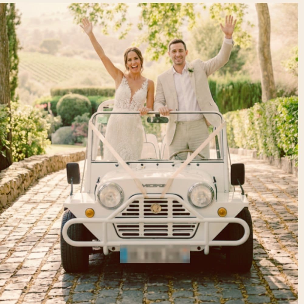 Mariés debout dans une Moke électrique blanche décorée de rubans, sur une allée pavée bordée de vignes