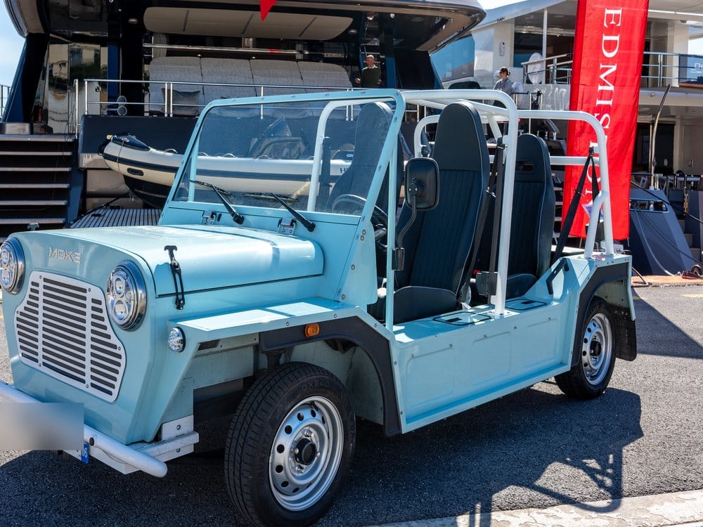 Moke électrique d'occasion Wave Blue de 2024, vue de trois quarts avant devant un yacht